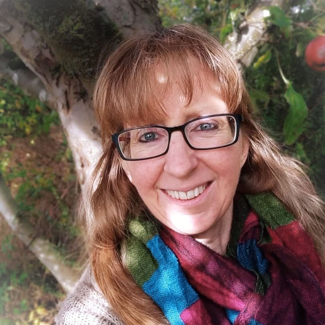 ich baum Barbara Mazur lächelt in die Kamera. Sie hat langes rotgoldenes Haar mit Ponyfrisur, trägt eine Brille.